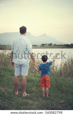 Father And Son Holding Hands Looking Out Over The Lake At The Mountain At Sunset