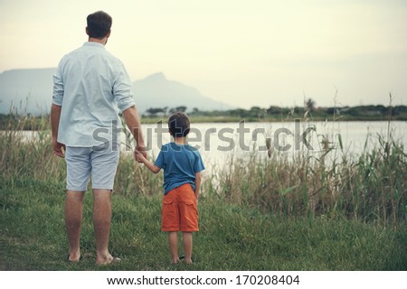 Father And Son Holding Hands Looking Out Over The Lake At The Mountain At Sunset