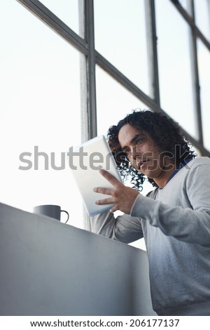 Mixed race man looking at his digital tablet with a hot beverage by the window