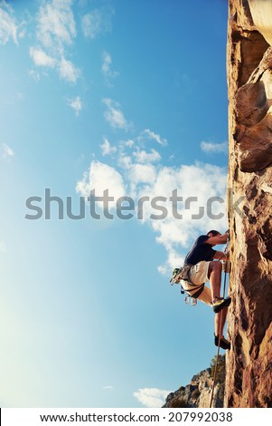 A man climbing up a really steep mountain attached to a harness and rope looking up against a blue sky in the outdoors