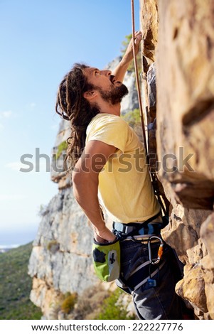 A man with dreadlocks climbing up a steep mountain with a harness and rope and looking up