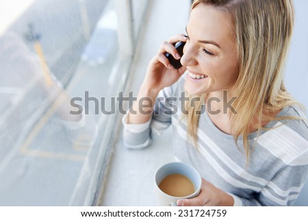 young woman chatting on phone while standing at the window and drinking coffee