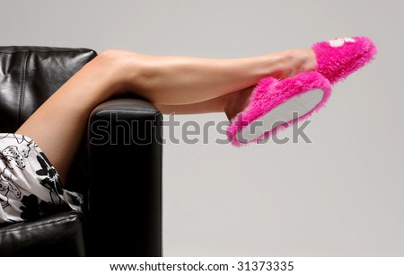 a girl wearing a white skirt relaxes and hangs her legs over the edge of a chair. she is wearing pink fluffy slippers.