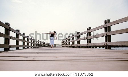 A girl walks alone on an abandoned pier balancing her way along the rotting beams. the sea breeze ruffles her white skirt and the wind plays in her hair.
