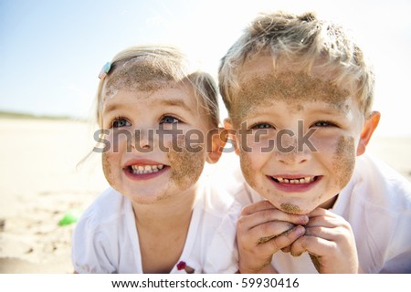 Brother and sister smile at the camera, brightly lit, on the beach, faces covered in sand.