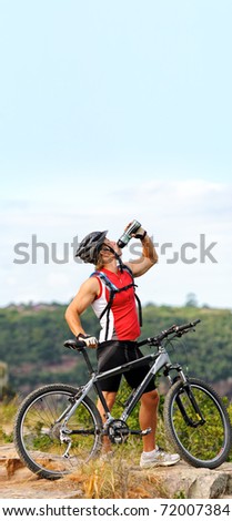 healthy young man stands triumphantly on a mountain and drinks water while holding mountain bike