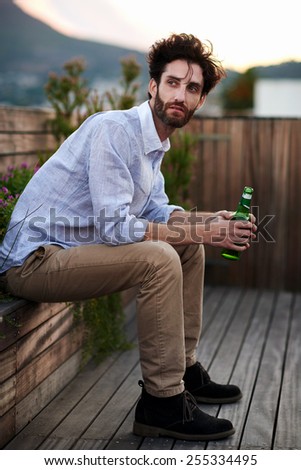 Portrait of young man holding bottle of beer sitting outdoors on rooftop terrace
