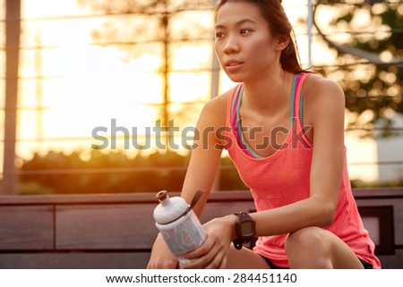sporty asian chinese woman sitting outdoors resting with water bottle after morning run