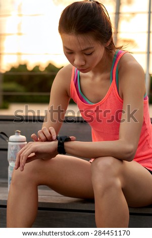 sporty asian chinese woman sitting outdoors checking workout time on wearable technology watch after run