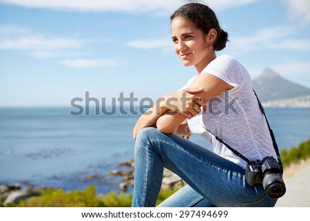 young, attractive female traveler sitting and enjoying the beautiful ocean view with digital camera hung on her shoulder
