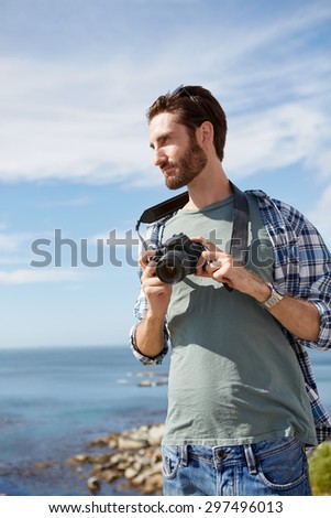young, attractive man stands near the ocean with digital camera in his hand