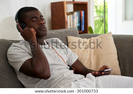 african black man listening to music relaxing on sofa couch with mobile cell phone in home living room