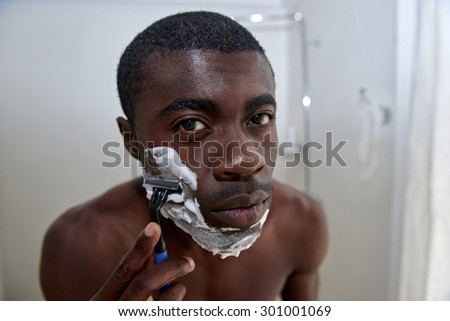 portrait of shirtless african black man shaving beard stubble face in mirror reflection for morning clean shaven look in home bathroom