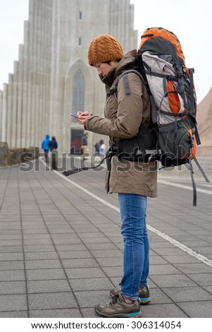 Young Asian woman in rain jacket and beanie carrying backpack busy with her mobile Phone in front of cathedral