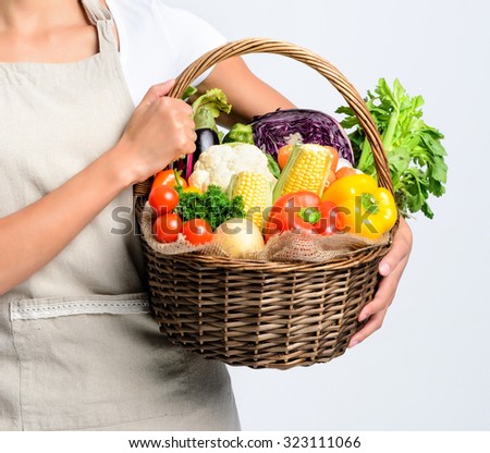 Anonymous young woman holding a basket full of fresh organic vegetable produce on grey background, promoting healthy diet and lifestyle