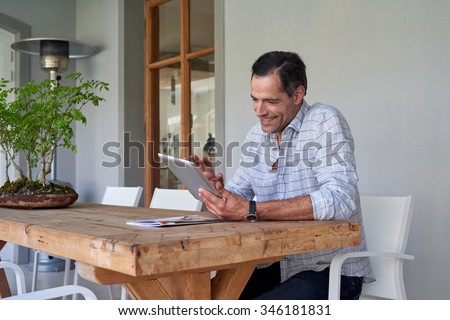 Handsome mature man sitting on his home patio looking up things on his tablet device