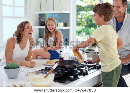 Happy caucasian family standing around stove, son making pancakes on stove
