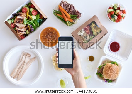 Hands holding smartphone mobile cellphone over different types of gourmet takeout, food delivery order app application concept