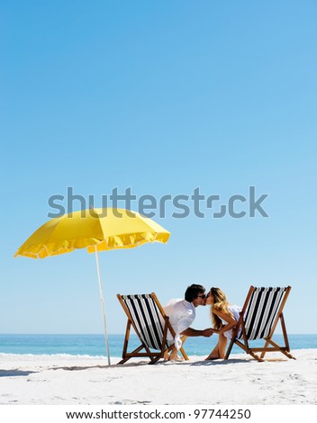 Beach summer couple kissing on island vacation holiday in the sun on their deck chairs under a yellow umbrella. Idyllic travel background.