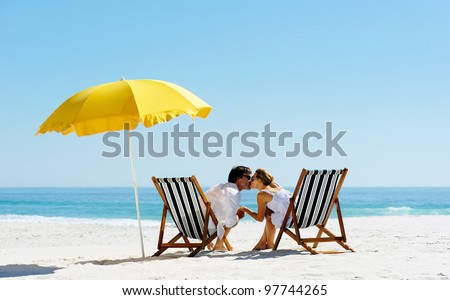 Beach summer couple kissing on island vacation holiday in the sun on their deck chairs under a yellow umbrella. Idyllic travel background.