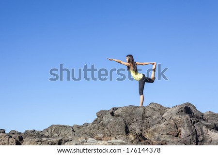 Young attractive woman doing dancers yoga pose on rocks against blue sky