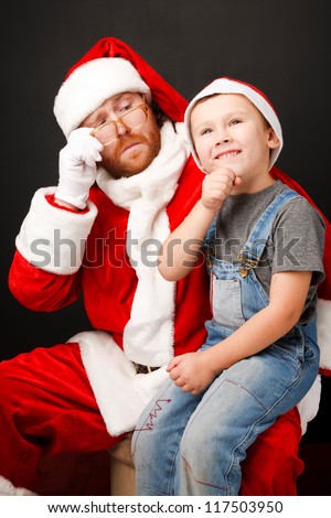 Boy is  sitting on Santas knee and asking about his  present.  Man,dressed in Santa costume looks tired and unhappy