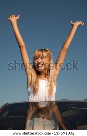 Cheerful young woman is standing in the  sport car with her arms raised