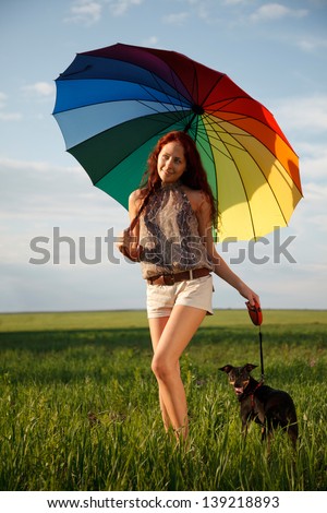 Young slim woman is walking on the field with the dog under big colorful umbrella