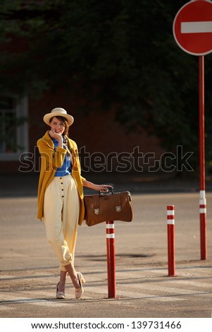 Young happy funny (vintage) dressed woman with retro suitcase stands on the street near red small columns.  Picture ideal for illustrating woman magazines.