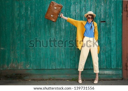 Young happy funny (vintage) dressed woman waves retro suitcase. Old green fence on the background.  Picture ideal for illustating woman magazines.