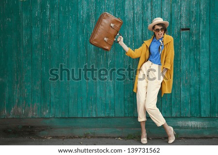 Young happy funny vintage dressed woman waves retro suitcase Old green fence on the background Picture ideal for illustating woman magazines Young happy funny vintage dressed woman waves retro suitcase Old green fence on the background Picture ideal for illustating woman magazines