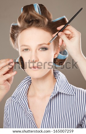 Young woman in curler in her hair and one eye with make-up.  With multiple hands applying make up.