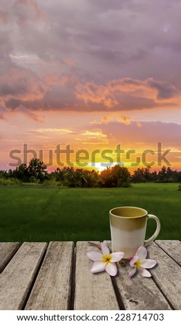 An image of coffee cup and flower on old wood table which have sunset as back ground