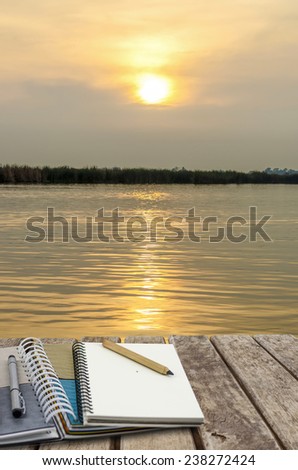 An image of note book, pencil on old wood table placed on the lake at sunset time