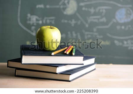 A school teacher\'s desk with stack of exercise books colored pencils and green apple. A green blackboard