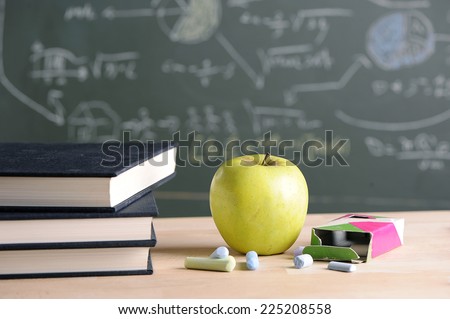 A school teacher\'s desk with stack of exercise books and apple in left frame. A blackboard in soft focus