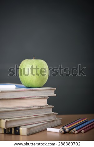 A school teacher's desk with stack of exercise books colored pencils and green apple. A green blackboard