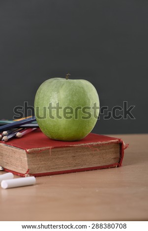 A school teacher\'s desk with stack of exercise books colored pencils and green apple. A green blackboard