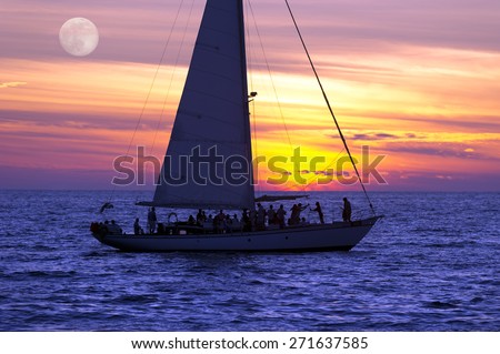 A sailboat full of people sails along the ocean at sunset as the moon rises in the background.