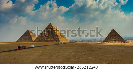 Cairo, Egypt - October 11, 2008: The three pyramids of Giza under a cloudy sky, with Cairo in the horizon wrapped by the mist and a group of vehicles driving through the road that crosses the plateau