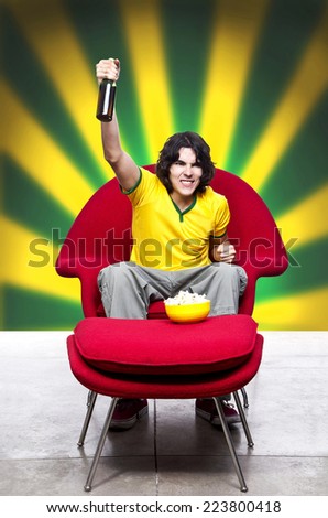Young Man with green  and yellow shirt or jersey  Shouting watching TV on a red sofa and a beer