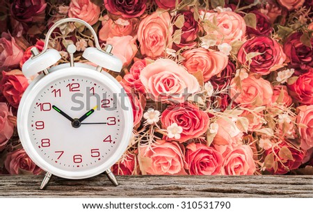 White alarm clock on wooden table with rose vintage flower background soft focus.