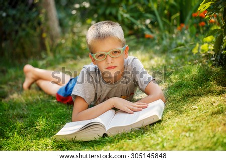 Cute baby boy with glasses lying on green grass, reading the book in summer day.