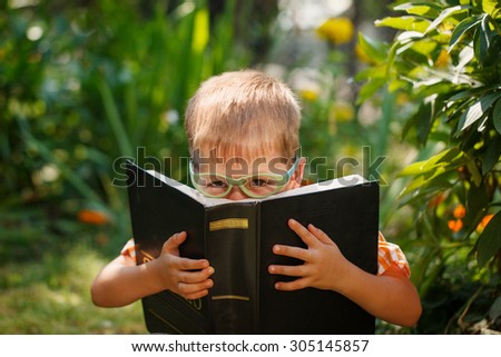 Beautiful kid boy with glasses, reading a book in garden, sitting on grass.