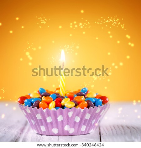 Decorated Birthday cupcake with one lit candle and colorful candies on yellow background. Holidays greeting card