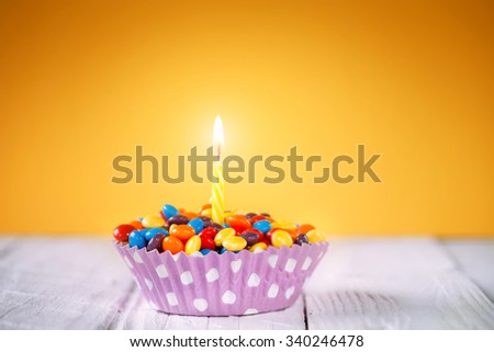 Decorated Birthday cupcake with one lit candle and colorful candies on yellow background Happy birthday card.