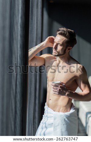 Handsome, muscular, young man drinking his morning coffee in a hotel room standing next to a window and looking against bright sunlight with towel wrapped around his waist
