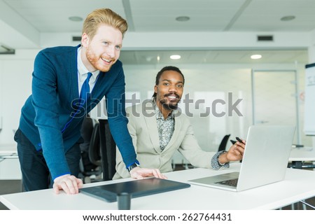 A black, young man with dreadlocks and a young, handsome  businessman discussing graphics design flaws in a nice white office