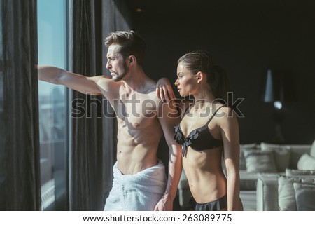 Athletic couple in love looking outside through the window of a luxurious hotel room and daydreaming