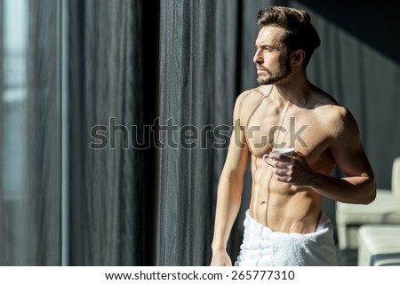 Handsome, muscular, young man drinking his morning coffee in a hotel room standing next to a window and looking against bright sunlight with towel wrapped around his waist